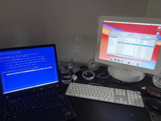 A desk with a Thinkpad T60 and a G4 iMac on it. The T60 is installing Windows XP and the iMac is running Mac OS 10.4. It’s running iTunes and pumping out some Deicide.