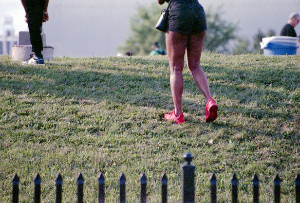 A woman in black shorts wearing bright red shoes climbs a grass hill to join other people. In the foreground, the top of an iron fence can be seen.