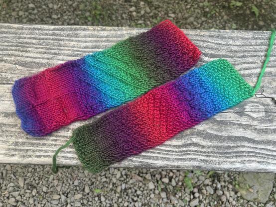 Two hand knit bookmarks on a wood bench. The yarn is self striping, slowly changing jewel tones. The bookmarks are knit in a variety of textures.