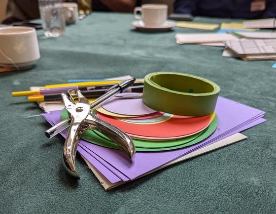 Tabletop covered in faux suede cloth. Pile of paper in multiple colours including green, purple and red. A roll of green masking tape. Hole punch and a pile of pens. Tea cups scattered across the table top.