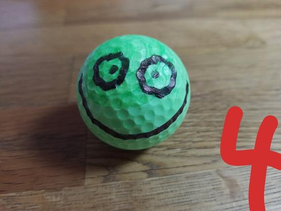 vor uns liegt ein grüner Golfball mit einem Smiliegesicht aufgemalt.
Zwei große runde Augen schauen uns freundlich an und ein breiter Grinsemund vervollständigt das freundliche Aussehen