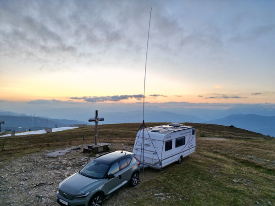 car and carvan with a long cb radio antenna beside a summit cross with mountains and clouds in the background