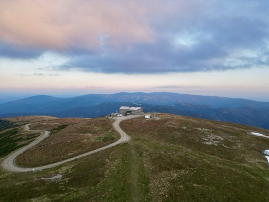 top of a mountain with a house and cars and a caravan with mountains and clouds in the background