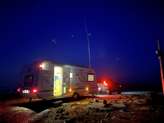 car and carvan with a long cb radio antenna beside a summit cross in the night with lights on