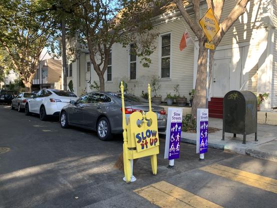 Slow Street with the two purple paddles announcing it. Next to them are deconstructed parts of a wooden chair, painted yellow, reassembled flat, with the message painted on: Slow it up