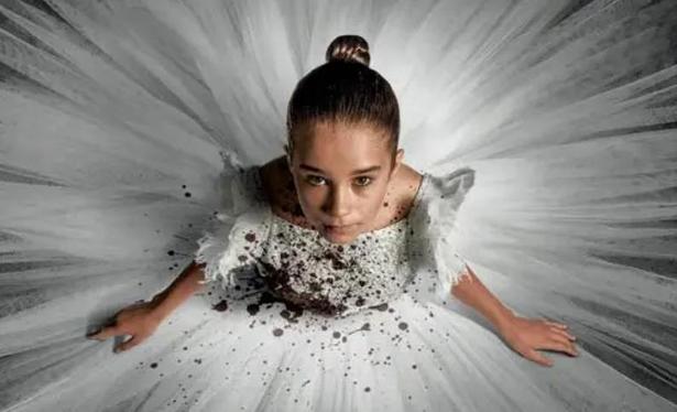 A little Anglo-Saxon girl with light brown hair in a tight bun wearing a ballerina dress splayed out around her like a white starburst as she looks up at the camera. There is mud spattered on the dress.
