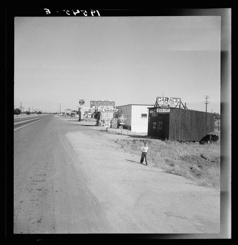 This black and white photograph captures a scene from U.S. Route 99 in California, specifically along the stretch between Tulare and Fresno where Highway City is located. The road appears wide and empty with a noticeable amount of tire marks indicating frequent use. On either side of the highway are various businesses including "BADMAN'S" which suggests an establishment possibly related to food or services for travelers.

To the right stands what looks like a large, weathered structure labeled as a garage, likely dealing in used cars given its sign, indicative of rural American roadside commerce from perhaps mid-20th century. The sky is clear with no visible clouds, and there's a stark contrast between the bright road ahead and the more shadowy area beside it.

A young man stands on the right side of the highway near this building; his posture relaxed yet attentive as he gazes towards something outside the frame. His clothing—dark trousers paired with a lighter shirt—is modest but practical, fitting for someone traversing through such an environment.

There's a sense of isolation and quietude in the image which is enhanced by the lack of any other visible activity or people on this stretch of road at what seems to be midday judging from the shadows cast. The overall composition provides a glimpse into rural life along America’s highways, portraying both its harshness—evid [...]
