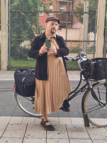 Selbstporträt in spiegelnder Scheibe. Ich sitze auf meinem Hollandrad mit einem Fuß auf dem Boden. Ich trage ein beigefarbenes langes Kleid mit plissiertem Rockteil und Hemdkragen, eine schwarze Jacke, schwarze Schiebermütze und schwarze klobige Spangenschuhe. Das Fahrrad ist ein "Damenrad" mit Schwanenhals-Oberrohr. Am Lenker ist ein Gepäckträger mit großer Bäckerkiste, am Hinterrad große Packtaschen.