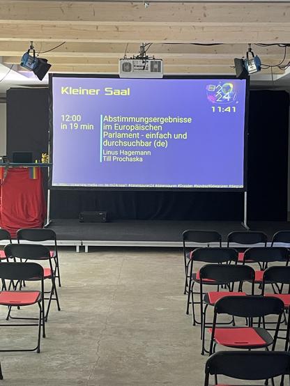 Foto des Veranstaltungsort, auf dem Beamer zu sehen ist der Zeitplan der Konferenz: Kleiner Saal, 12:00 Uhr, Abstimmungsergebnisse im Europäischen Parlament - einfach und durchsuchbar (de)