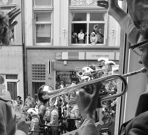 das schwarzweißbild zeigt aus einem fenster aufgenommen einen karnevalsumzug.
im fenster rechts steht ein karnevalist, spielt trompete und winkt auf die straße. dort fährt gerade ein musikwagen, ein posaunenchor vorbei. einige sind irritiert und schauen in das fenster. links winkt eine weitere person aus dem fenster und hält ein kölsch in der hand.
auf der gegenüberliegenden straßenseite stehen in einem anderem fenster ebenso verkleidete karnevalisten und feiern den karnevalsumzug im viertel.