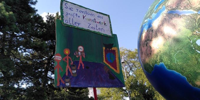 Demoplakat. Text: "Sie zerstören das größte Kunstwerk aller Zeiten"

Darunter ein gemaltes Bild von einem Museum, wo ein Bild der Erde hängt. Daneben drei Personen, gelabelled mit "Exon", "Shell" und "OMV" die ÖL auf das Gemälde spritzen.