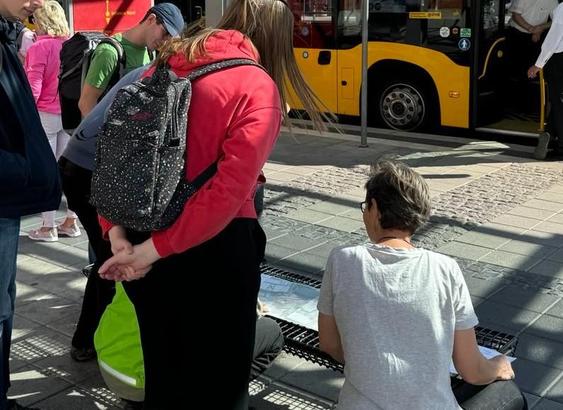 Menschen hocken vor den aufgefalteten Plänen an einer Sitzbank vor dem Terminal.