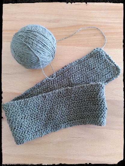 Ein im Rosenmuster gehäkeltes, schräg über sich selbst gelegtes Stirnband, der Rand ist in Krebsmaschen, darüber das zugehörige, runde Garnknäuel. Farbe ist ein khakigrün.