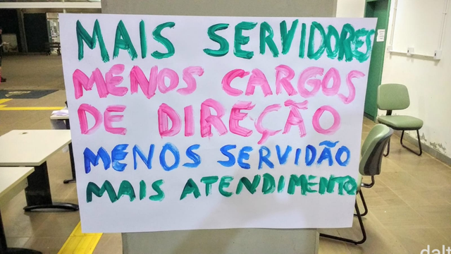 Cartaz, feito de uma folha branca de cartolina e letras maiúsculas coloridas pintadas com tinha guache, sendo cada frase com uma cor, colado em uma porta ou parede de vidro:

Mais servidores
Menos cargos de direção
Menos servidão
Mais atendimento