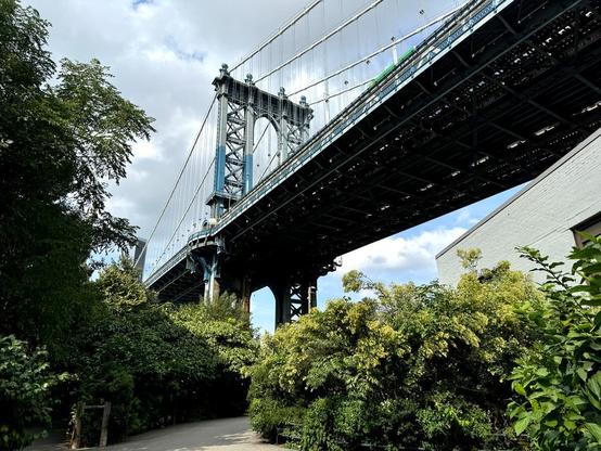 A close view of the iconic Manhattan Bridge from a park walkway just below the bridge. The span of the bridge juts diagonally across the frame and is set against blue skies with a mix of white and gray clouds.
