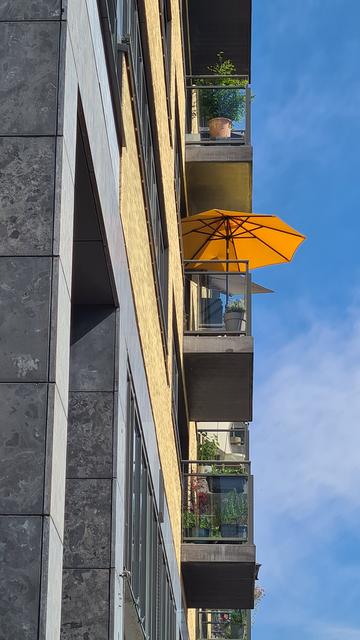 Three balconies consisting of a concrete slab with glass siding above one another as seen from the side looking up from street level on a sunny day. All three have one or more potted plants. The middle one also features a bright orange parasol.