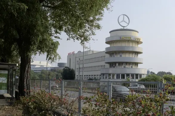 The old AVUS control tower, now motel. A four-story round tower with circular terraces, attached to a rectangular building behind it. The top terrace has a MOTEL AVUS sign (M is damaged) and there is a massive Mercedes star on top. The building is photographed across the Autobahn, with road fencing, some green plants and trees, and part of a bus stop in the foreground.