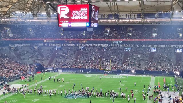 Stadion. Auf der mehrseitigen Anzeige wird der Sieger des Spiels angezeigt. Unten klatschen sich die Spieler zum Spielende in der Mitte des Platzes ab.