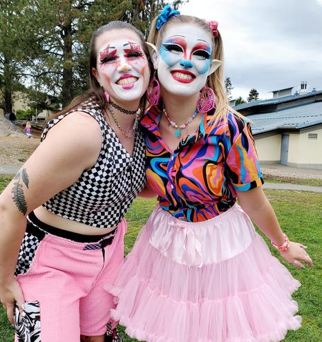 Two members of clown car drag show troupe, grinning at camera. Wearing harlequin clown makeup. Dressed in bright costumes.