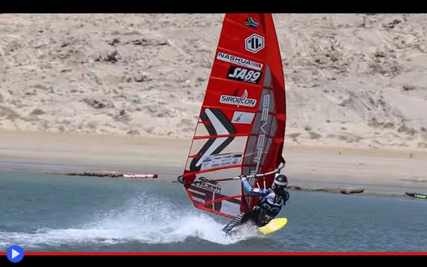 Surfista su una tavola a vela si regge saldamente mentre accelera all'interno di un canale artificiale. Sullo sfondo è visibile un dirupo sabbioso totalmente privo di vegetazione.