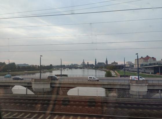 Ausblick aus einem Zugfenster. Schienen und eine Straße mit Autos überbrücken den Fluss Elbe. Im Hintergrund ist klein die Dresdner Altstadt zu sehen.