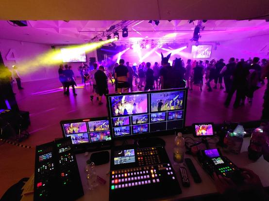 Picture of the dead dog party, taken from behind the video mixing desk.
In the background, people are dancing on the floor, humans and fursuiters alike.
In the foreground are monitors with the different camera views and consoles for selecting cameras, controlling their settings and also a PTZ joystick.