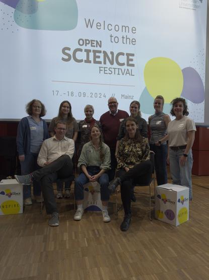 A group of people from different consortias sitting and standing together in front of a screen with the title "Welcome to the Open Science Festival, 17.–18.09.2024, Mainz"