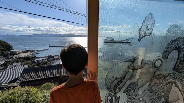 Ogijima Pavilion's main window. The three windows panels are open and merged into one drawing: an octopus grabbing a fish and more (sorry, it's impossible to describe). In the background, some roofs from Ogi village and the Seto Inland Sea. In the center of the picture, a little boy seen from behind is looking at the sea.