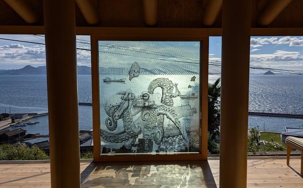 Ogijima Pavilion's main window. The three windows panels are open and merged into one drawing: an octopus grabbing a fish and more (sorry, it's impossible to describe). In the background, some roofs from Ogi village and the Seto Inland Sea.