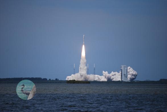 An outdoor, daylight photograph of a rocket launching from the other side of a body of water. The rocket, copper-colored on the bottom with a white, cynlindrical middle and rounded top.
A column of bright orange flame emerges from the bottom of the rocket and a trail of dust and smoke lead from the rocket down to its pad on the horizon.