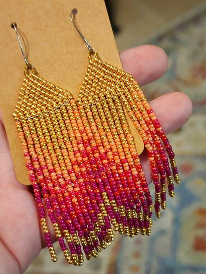My hand holding a pair of Cvkvlv beadwork Totkv Lane Chevron Brick Stitch Fringe Beaded Earrings. Tiny beads in gold, salmon, magenta and fuchsia colors.