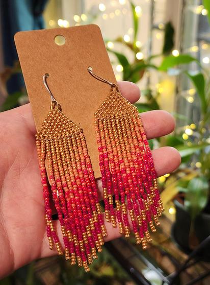 My hand holding a pair of Cvkvlv beadwork Totkv Lane Chevron Brick Stitch Fringe Beaded Earrings. Tiny beads in gold, salmon, magenta and fuchsia colors this time under more artificial light showing the beads under different light conditions.