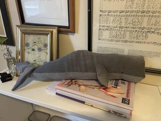 A sperm whale (long, square nosed with short flippers) made of grey denim. She is about 2 feet long and has small, embroidered eyes.