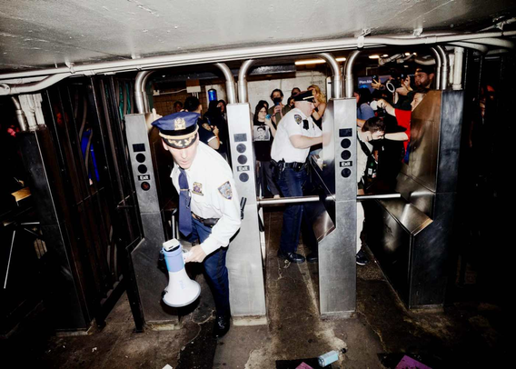More than a hundred people evaded fare by hopping the turnstiles en masse at the 8th Street entrance of West 4th Street Station. In this photograph, police are seen flailing around, with one officers brandishing a bullhorn, as they try to seize control of the turnstiles.