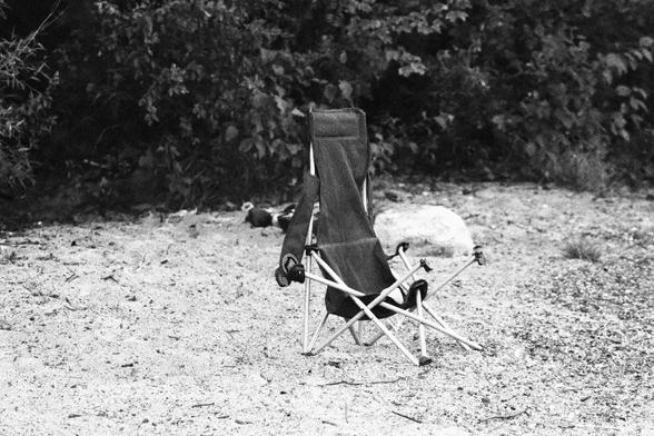 An abandoned folding chair sits on the coarse sand of the beach, behind it can be seen a large rock, well further back is dense bush - a medium shot.