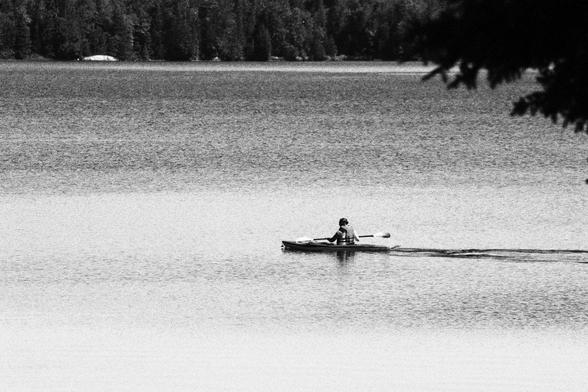 A Long shot of a lone kayaker making their way across the water. In the far background is the north shore of Elliot Lake. Dense coniferous trees hug the shore.