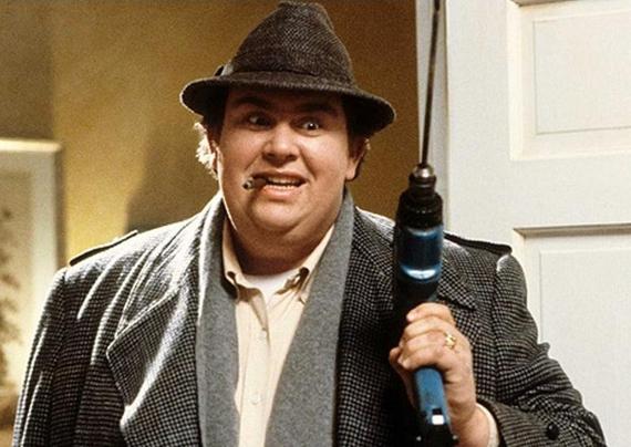John Candy wearing a woolen overcoat, Trilby hat, cheroot in his mouth, holding a drill in his hand while he has a crazed look on his face.