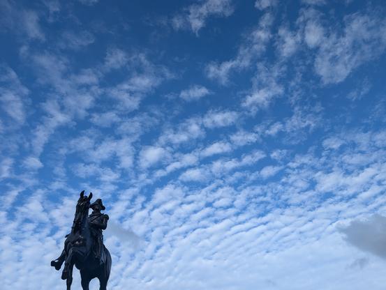A bronze statue of a horsed George Washington brandishing a sword, posed against a vast blue sky threaded with dramatic clouds.