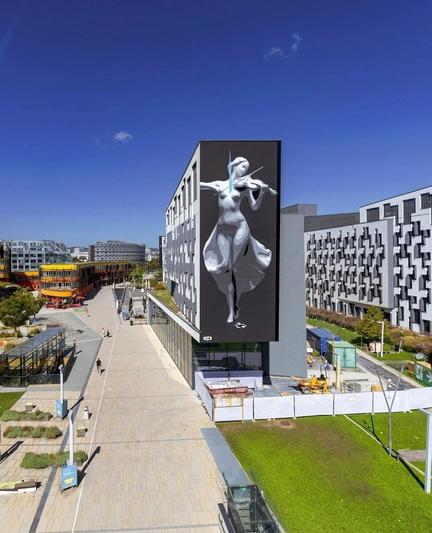 Streetartwall. The portrait of a young woman playing the violin was sprayed/painted on the outside of a modern, four-storey building. The mural is painted entirely in black, gray and white. The delicate female figure wears a long, flowing white dress and looks like a porcelain figurine. She gracefully holds a violin in her hands and plays. In front of her are coins as a reward for her art and a small blue line through the picture marks the signature of the artists Inoexpo. The background is dark gray and makes the huge painting stand out even more. (The futuristic surroundings in the photo is a university campus with long, wide paths and other high-rise buildings.)