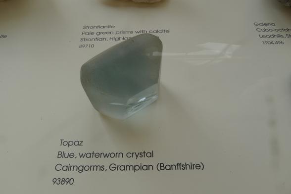 a clear pale blue crystal sits on the white card base of a museum display case. Beside the crystal, on the base, are printed the words "Topaz. Blue, waterworn crystal, Cairngorms, GRampian (Banffshire) 93890"