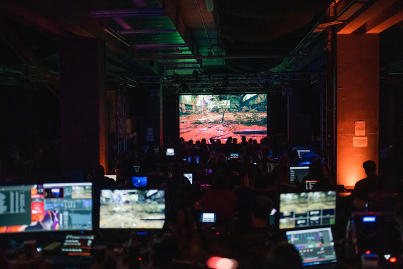 Picture taken from the FOH desk showing the main hall of Deadline at the Orwohaus. An entry in the animation competition is playing on the bigscreen, with many people gathering in front of it to watch.
