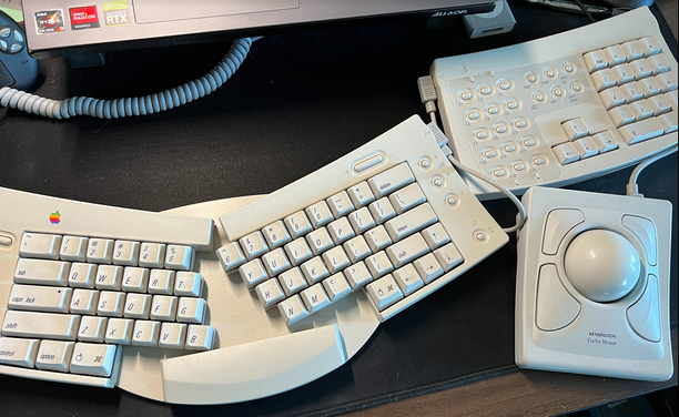 A desk with several Apple Desktop Bus (ADP) items: an Apple Adjustable Keyboard (M1242) rotated into an ergonomic angle with an attached number keypad with arrows and function keys, and a Kensington Turbo Mouse trackball. All items and cords are late-90s grey-beige.