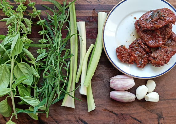 Auf einem Brett noch ungeschnitten Basilikum, Estragon, Stangensellerie, Knoblauch, Schalotten + getrocknete Tomaten.