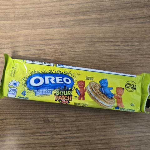 Photograph of a sleeve of a 4-pack of Oreos Sour Patch Kids flavored cookies on a wooden surface. The cover has the logos on the left and on the right is a vanilla wafer cookie with a white cream filling with colored speckles throughout. Around that are three Sour Patch Kid candies being naughty with cans of spray paint.