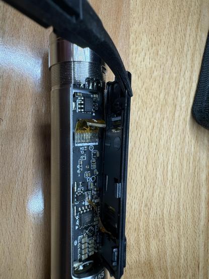 A close up photo of a partially opened electric screwdriver. A tiny zif style ribbon cable is visible connected to the PCB but crucially disconnected from the display that is being held aloft by tweezers. The screwdriver has a metallic cylinder look to it and it's resting on a wooden workbench.