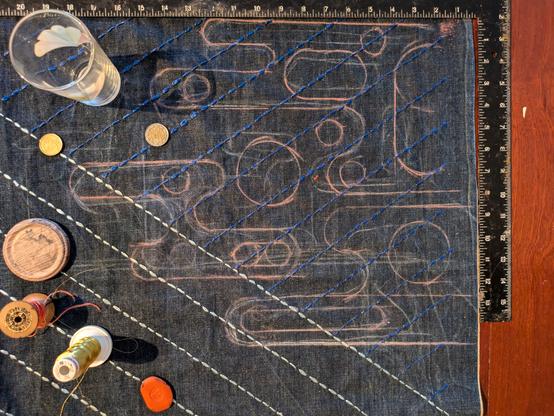 Part of a denim sashiko quilt cover in design phase. Diagonal pale blue and deep blue lines are overlaid with rough abstract curved forms in tailors chalk. Also visible are bobbins of thread and some round objects used to draw the curves