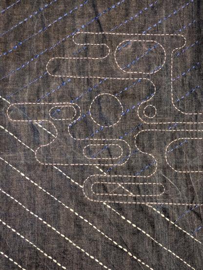Blue denim fabric stitched with sashiko in pale blue and deep blue opposing diagonals. The design from the previous image is now stitched in a pale blue-grey silk. It is semi-circles connected by straight lines, making abstract unconnected shapes