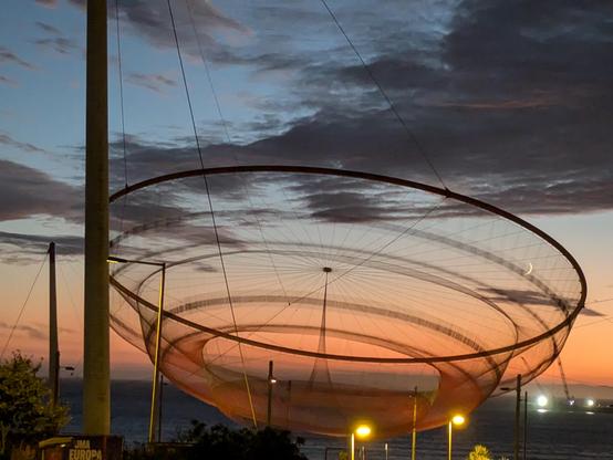 A view of She Changes, a massive installation by Janet Echelman. It looks like a huge circular net suspended against a late evening sky and the Atlantic Ocean, full of soft colours from dusky orange to a deep grey blue. There are layers of gentle soft clouds which have hints of purple in their depths