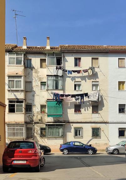 Fachada de un edificio con signos de deterioro. En los dos pisos superiores hay cuerdas de ropa en cada con ropa oscura tendida. En la calle se ven aparcados varios coches, uno de ellos con el capó abierto.