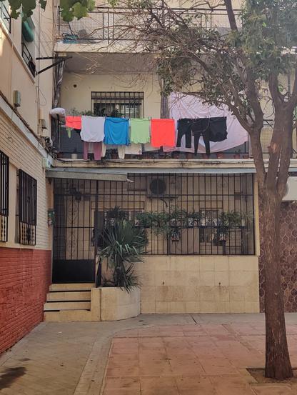Ropa de deporte de colores fluorescentes tendida en un balcón de una casa de varios pisos. La entrada, en el piso de abajo, está cerrada con una reja. Delante de la fachada hay un árbol.